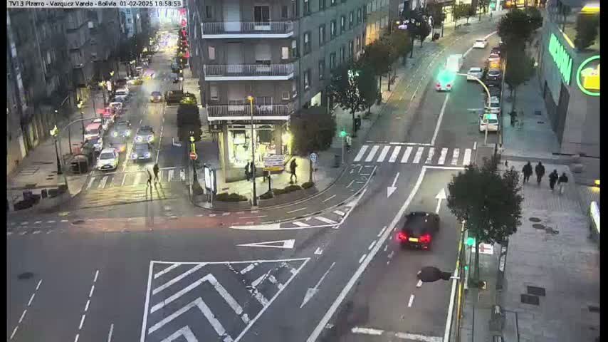 An aerial view of a city intersection at night, showing several cars and pedestrians, with streetlights illuminating the scene under twilight conditions.