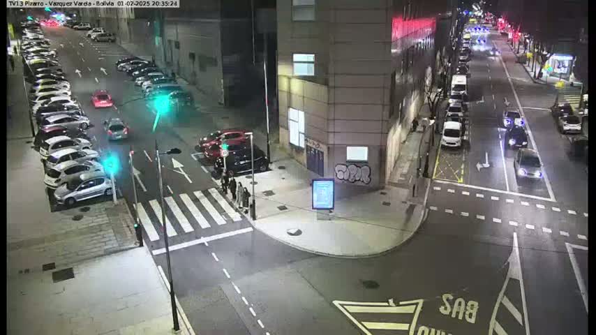 A nighttime street scene shows several cars parked and driving on a city street, intersecting with a crosswalk where a few pedestrians are walking.