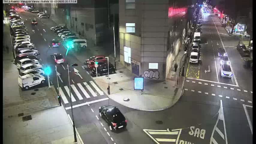 A nighttime street scene shows several cars parked along the sides of a road, with a few cars driving on the road and a crosswalk, under clear skies.