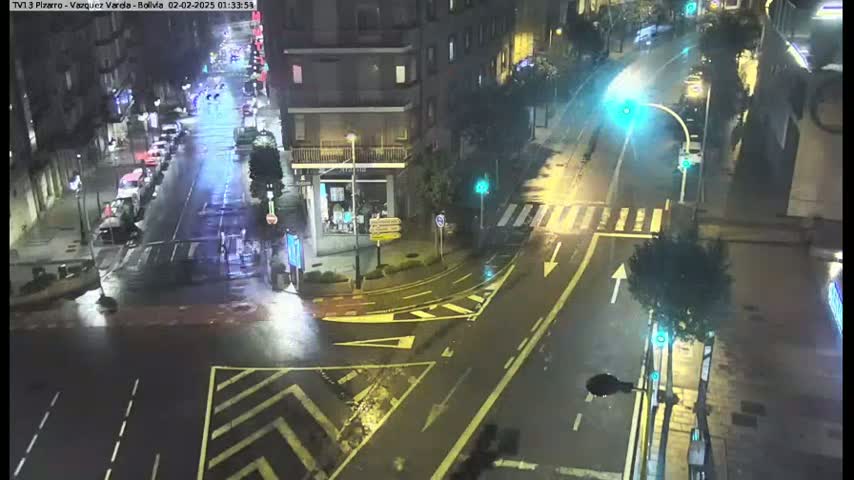 A rain-slicked city intersection at night is sparsely populated with vehicles and streetlights illuminating the wet roads.