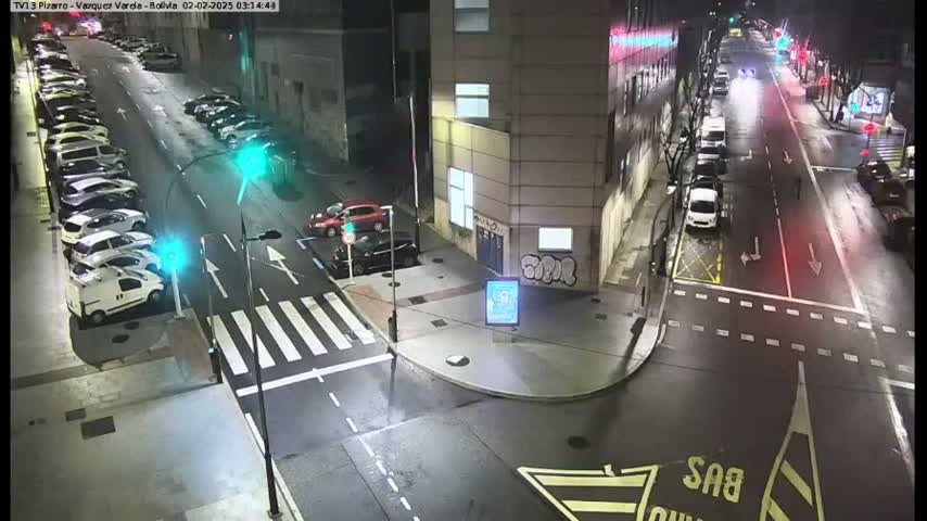 A high-angle night view shows a city intersection with cars parked along the sides of the street and a few cars driving.