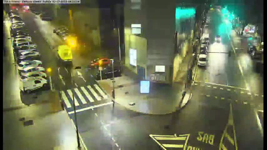 A nighttime, wet street scene shows several parked cars, a few moving vehicles, and a crosswalk, all viewed from above.