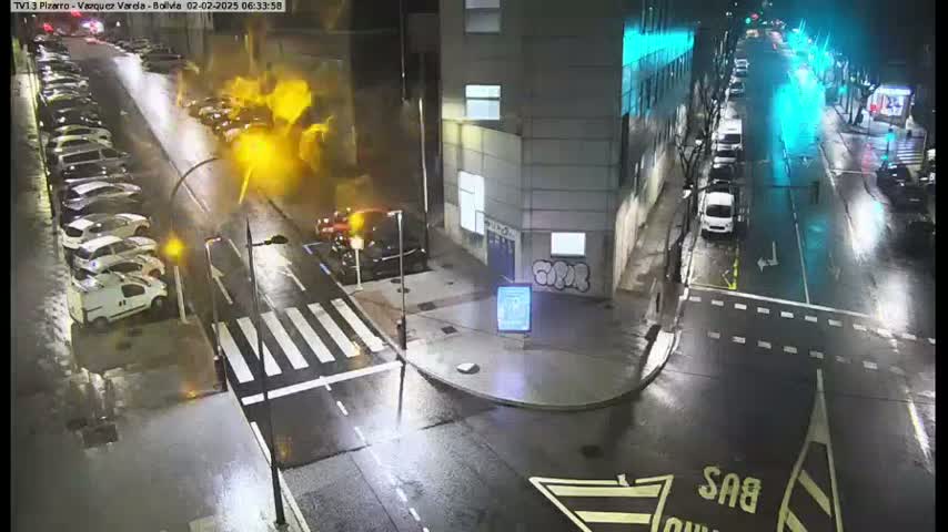 A wet, nighttime street scene shows cars parked along the side of a street and driving down the road, with a building visible in the background.