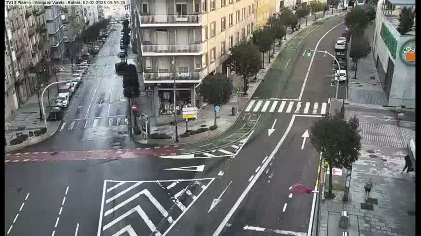 A wet city street intersection shows several cars parked along the sides of the roads, with a few cars traveling through the intersection.