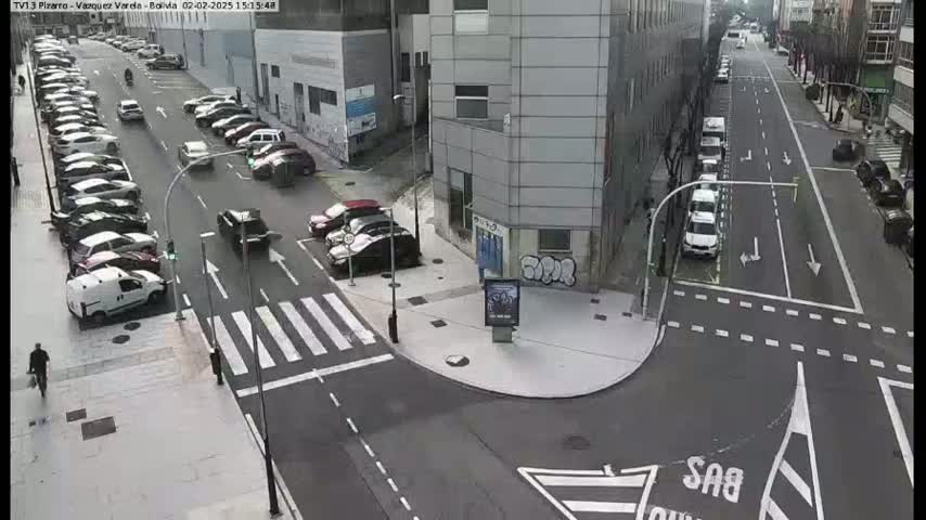 A street intersection with parked cars lining both sides of the road, a few cars driving, and a pedestrian walking.