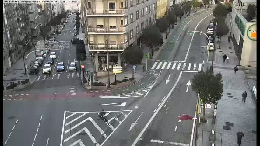 A high-angle view of a city street intersection with several cars and pedestrians, on a partly cloudy day.