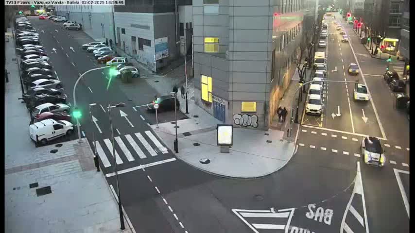 A street intersection with parked cars along the side and several cars driving on the roads in the evening.