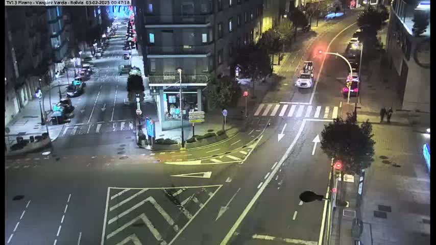 A nighttime, wet street intersection shows several cars and pedestrians, with buildings lining the streets.