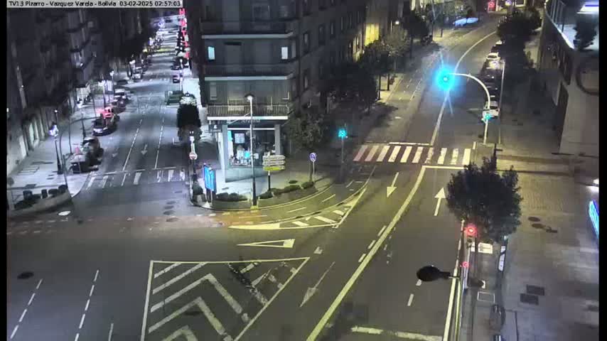 A nighttime street intersection shows several cars parked along the sides of the road and a few cars driving through the intersection.