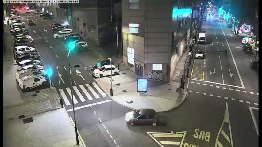 A nighttime street scene shows several parked cars and a few cars driving on a road that intersects with another road, with traffic signals visible.