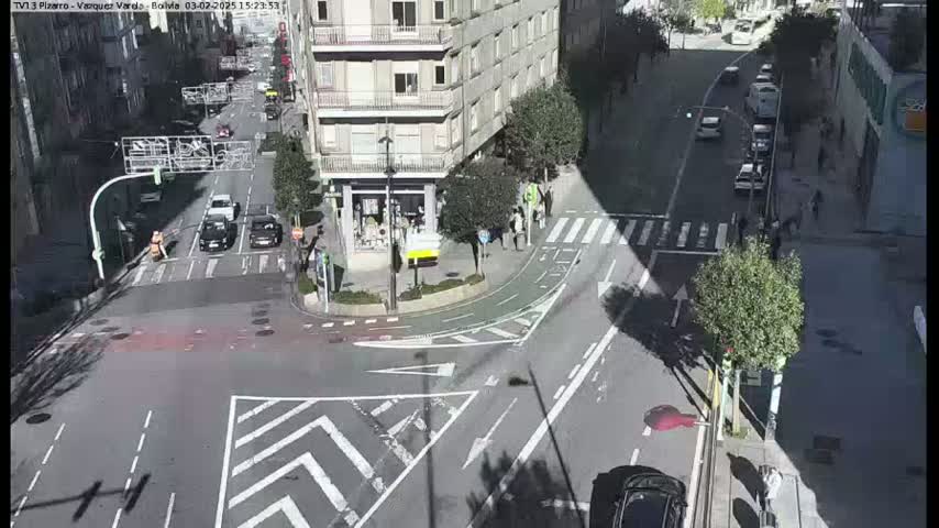 A sunny city street intersection shows several cars and pedestrians, with a building visible in the background.
