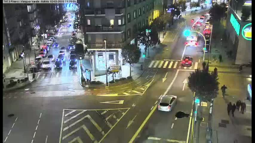 A wet city intersection at night, with cars driving on roads lined with buildings and pedestrians walking on sidewalks.