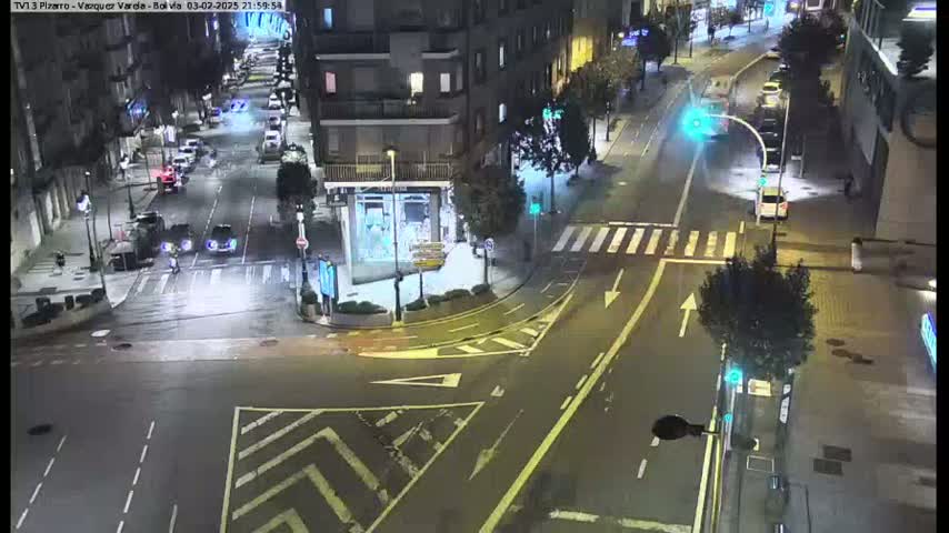A nighttime street intersection shows several cars and pedestrians moving through a wet, city street.