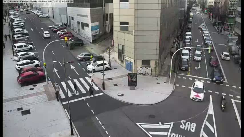 A city intersection with parked and moving cars, pedestrians crossing a crosswalk, and buildings in the background on a mostly sunny day.
