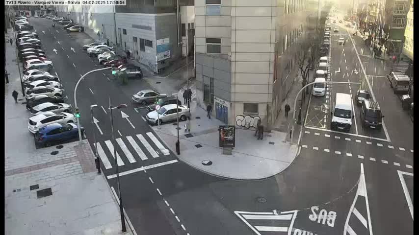 A sunny city intersection shows several cars parked along the sides of the street and others driving through, with pedestrians walking near the corner of a building.