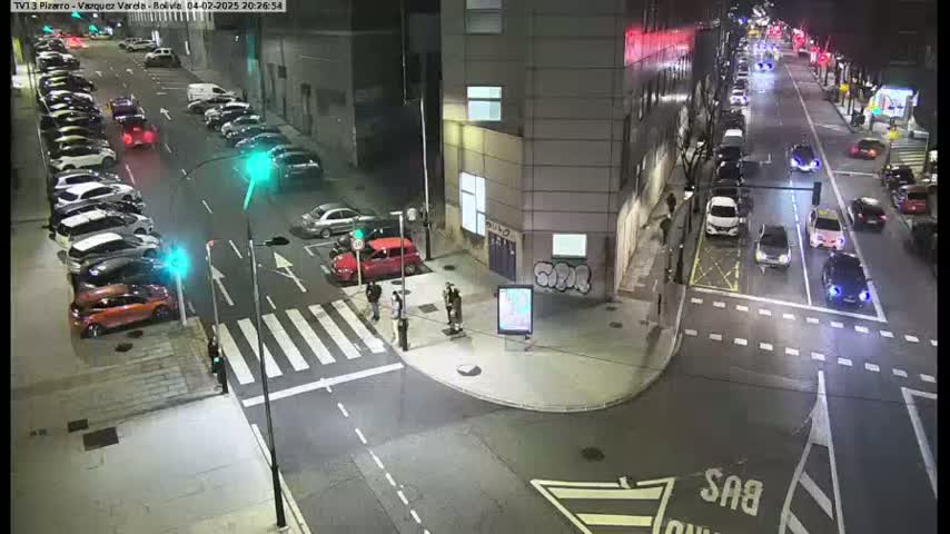 A nighttime street intersection shows several cars parked along the sides of the street and others driving through, with a few pedestrians standing near a crosswalk.