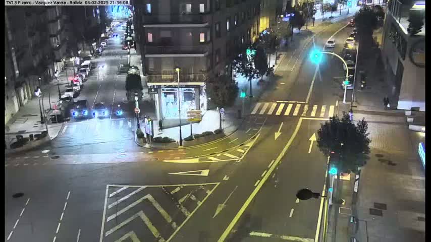 A wet, nighttime street intersection shows several cars and a crosswalk, with buildings and streetlights visible in the background.
