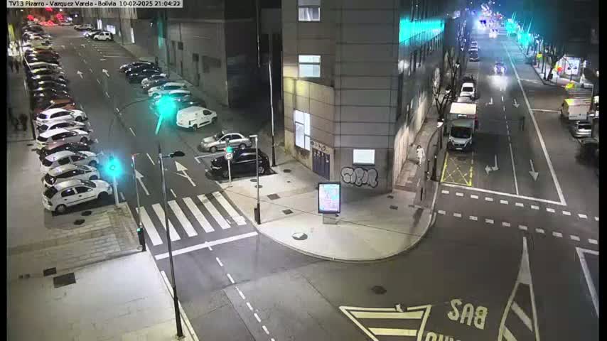 At night, a street intersection shows several parked cars along the side of a building and a few cars moving along the road.