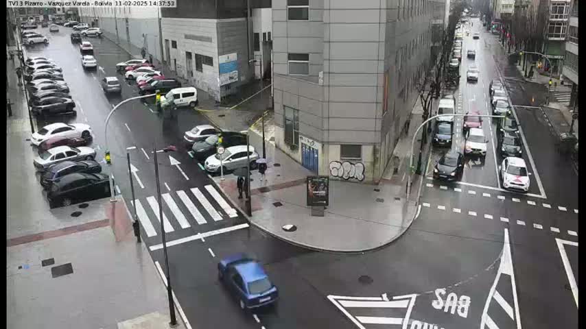 A rainy city intersection shows several cars parked along the street and several more driving through the intersection, with pedestrians near a crosswalk and a building in the background.