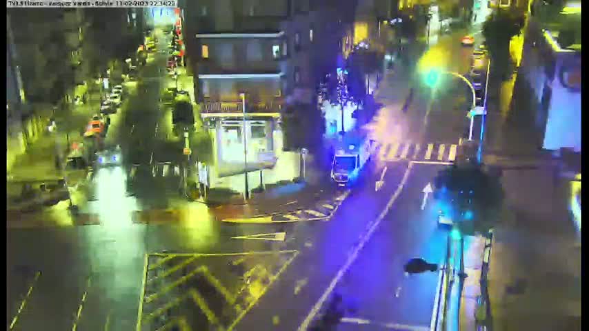 A nighttime, rainy scene shows an intersection with traffic, including an emergency vehicle with flashing lights, and buildings in the background.