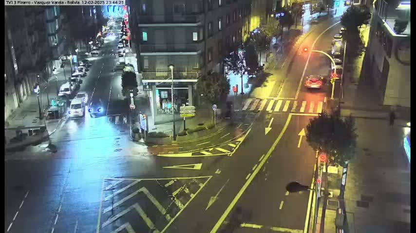 A rain-slicked city intersection at night shows several cars navigating a multi-lane roadway, with buildings visible on either side.