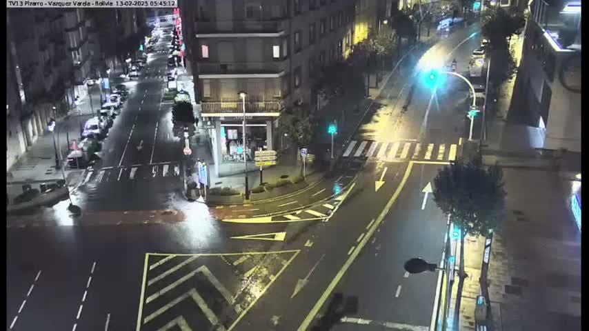 A wet, nighttime street intersection shows cars parked along the sides of the roads and a few driving through the intersection.