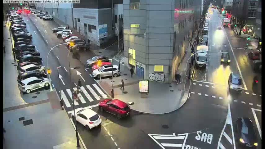 A wet city street scene shows cars parked along the side of a street and cars driving down the street, with pedestrians crossing at crosswalks near a modern building.
