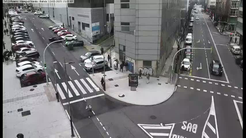 An overhead view of a city street intersection with parked cars along the sides, several pedestrians near a building, and cars driving through the intersection on a cloudy day.