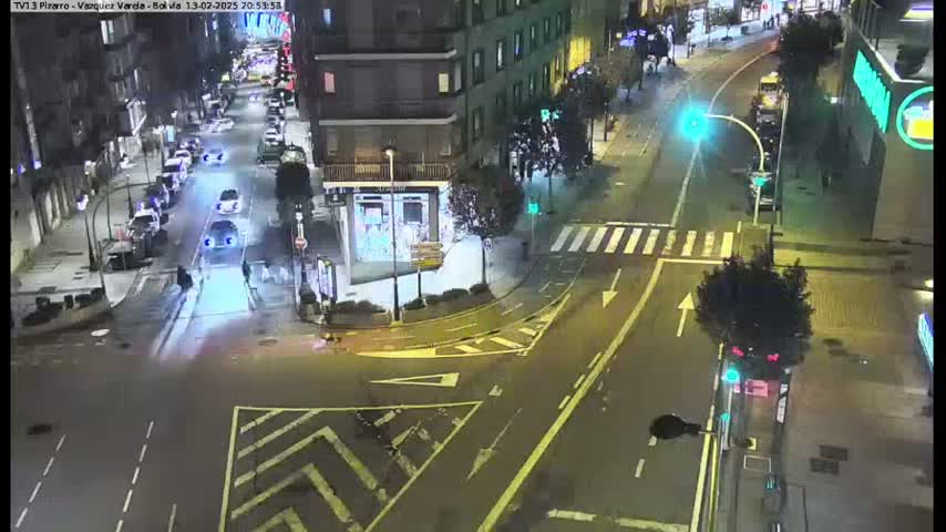 A nighttime street intersection shows several cars and pedestrians, with traffic lights and crosswalks visible under dark, clear skies.