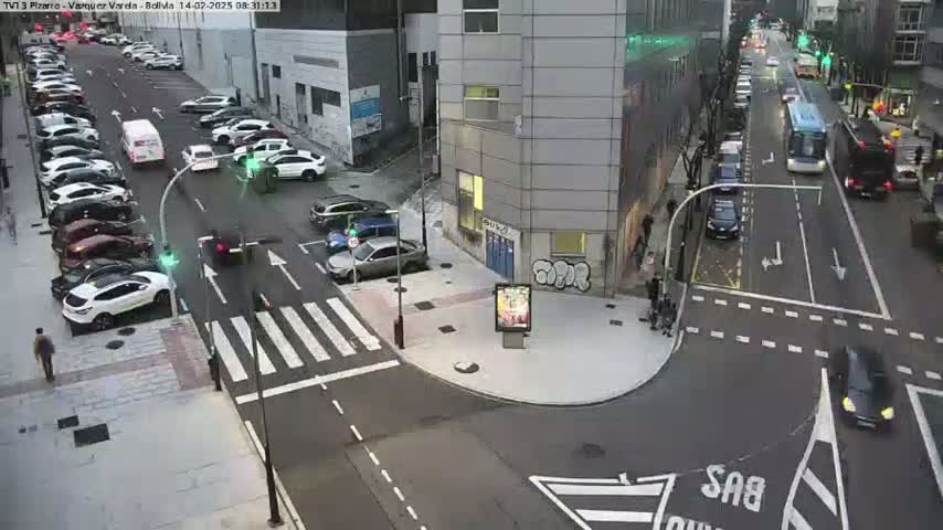 A city street intersection with parked cars lining the sides of the road and several vehicles in motion, under overcast daytime conditions.