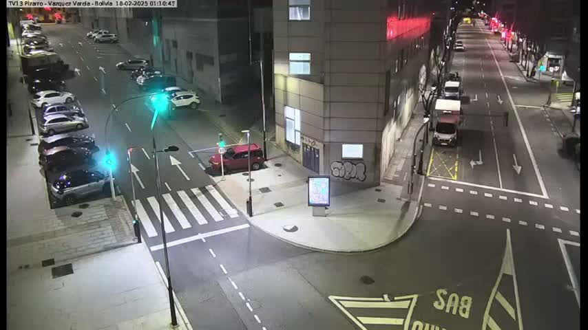 A nighttime street intersection with cars parked along the sides and a few vehicles driving on the roads, illuminated by streetlights.