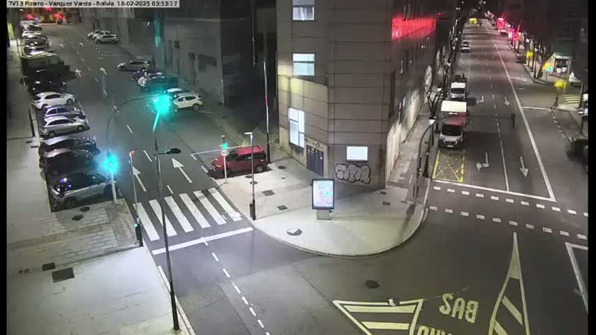 A nighttime street scene shows parked cars along the side of a road that intersects with another road, with a few cars driving and traffic signals present.