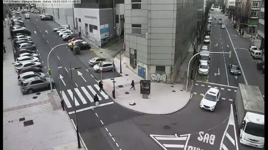 A city street intersection with pedestrians crossing a crosswalk, several parked cars along the sides of the road, and cars driving on the road, under an overcast sky.