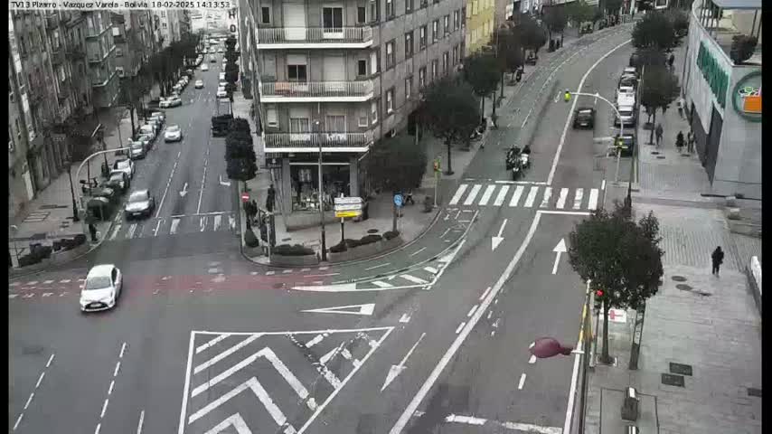 A city street intersection with several cars and pedestrians, under an overcast sky.