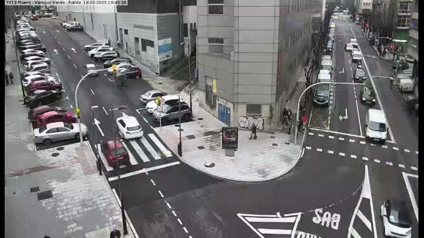 A city street intersection with parked cars lining the sides, several vehicles in motion, and a few pedestrians visible on a cloudy day.