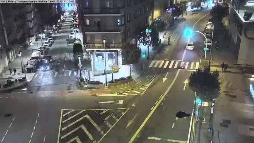 A nighttime aerial view of a city intersection with several cars and pedestrians, under wet conditions.