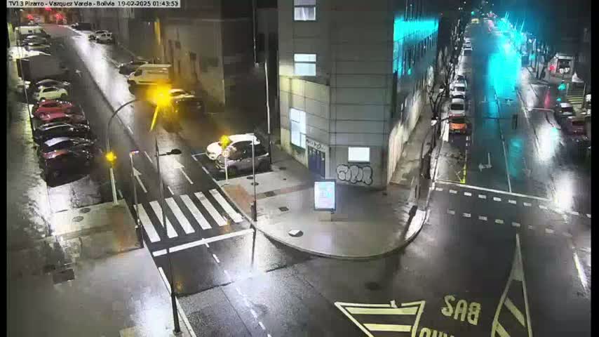 A wet, nighttime street intersection shows several parked and moving cars around a building.