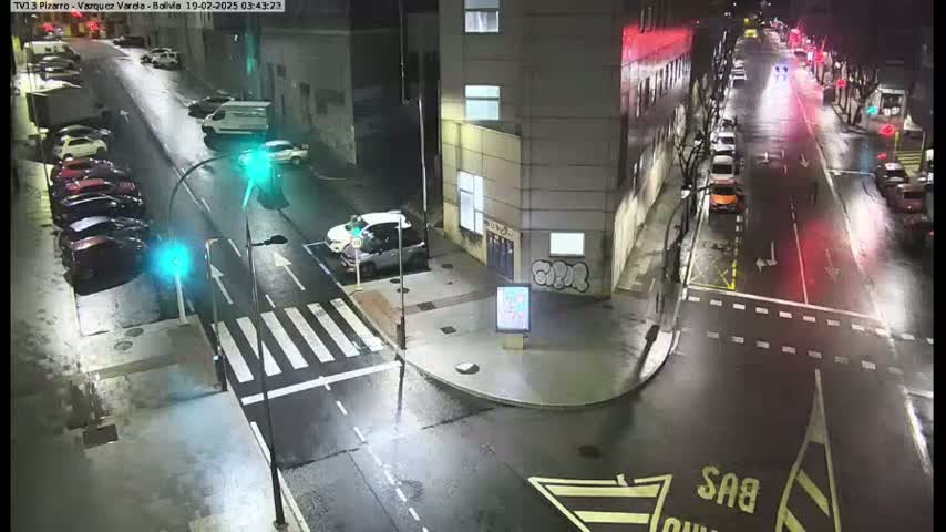 A wet, night-time street scene shows parked cars lining a street and several cars driving down a street that intersects with it, all near a multi-story building.