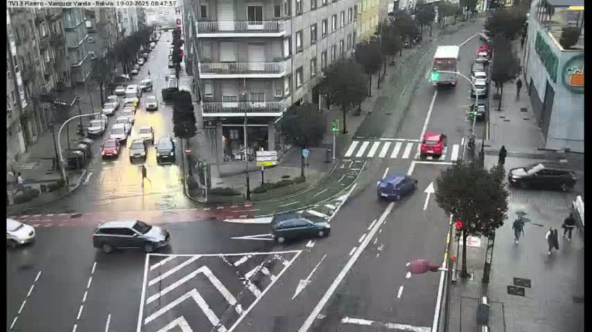 A rainy city intersection shows several cars navigating a crosswalk and streets, with pedestrians walking on the sidewalks.