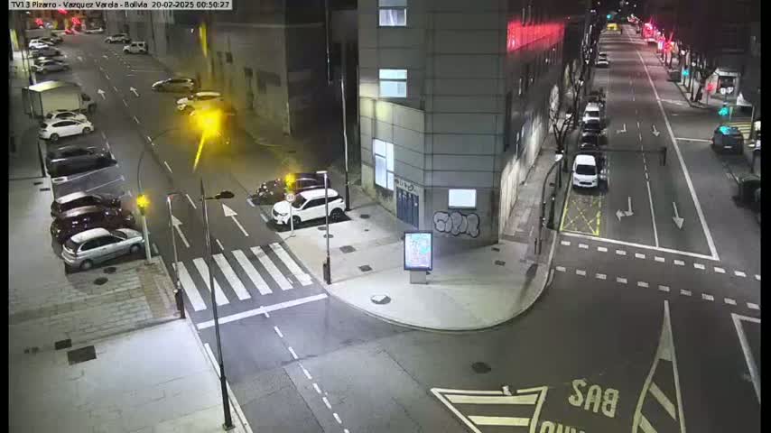 A nighttime street scene shows several cars parked along the curb and driving on a multi-lane road that intersects with a crosswalk.