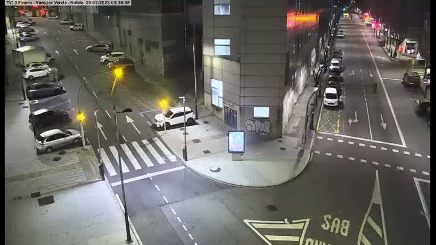 A nighttime street scene shows several cars parked and driving on a multi-lane road that intersects with another road, near a building with graffiti.