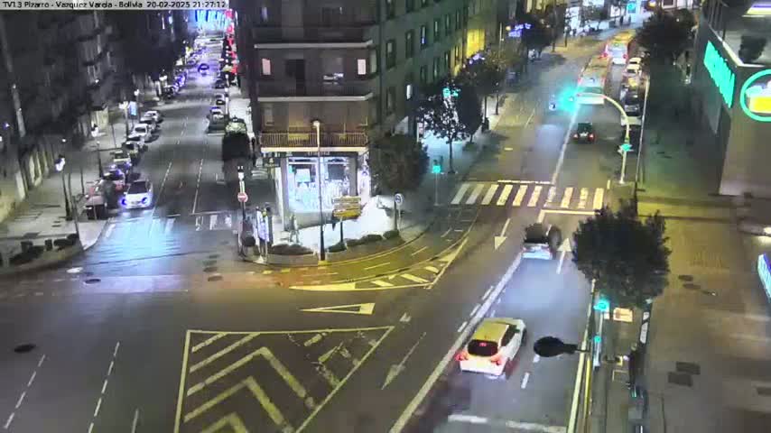 A nighttime aerial view of a city street intersection with several cars and a few pedestrians, showing moderate traffic under clear skies.