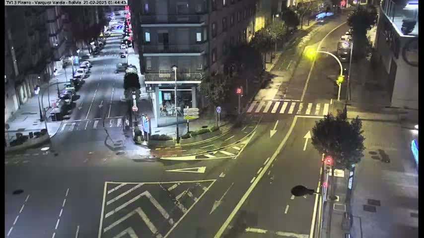 A nighttime aerial view of a city intersection with several cars parked along the streets, some cars driving through the intersection, and trees lining the roads.