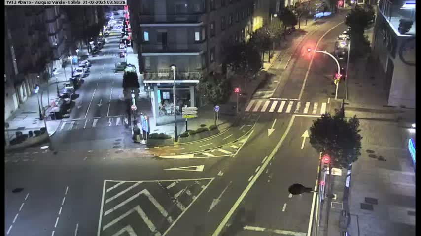 A nighttime aerial view of a city intersection with cars parked along the sides of the street and a few cars driving through the intersection.