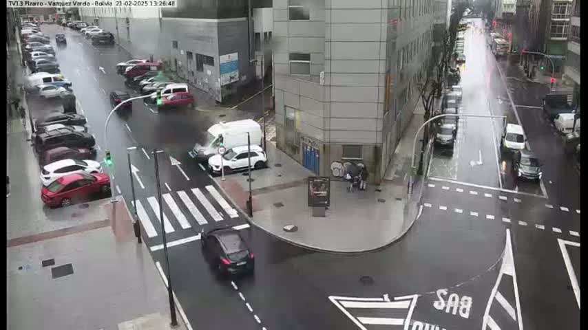 A rainy city intersection shows several cars parked along the sides of the street and others driving through the intersection, with a few pedestrians walking near a building.