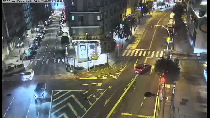 A nighttime, wet city street intersection shows several cars driving through a crosswalk and turning at the corner, with buildings lining the street.