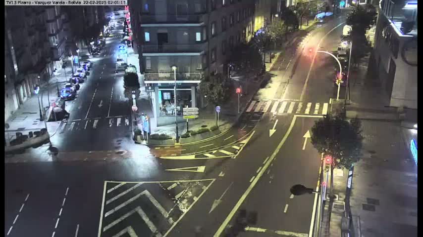 A wet, nighttime street intersection shows several parked cars lining the streets and a few vehicles on the road.