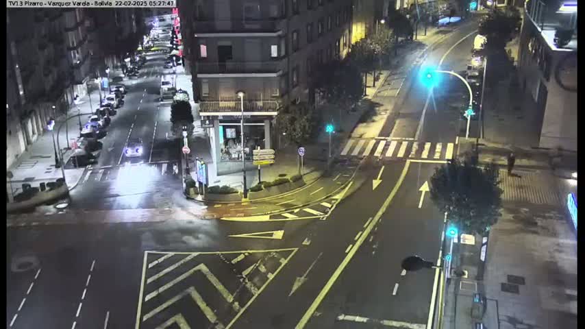 A wet, nighttime street intersection with cars parked along the sides of the road and a few cars driving through the intersection.