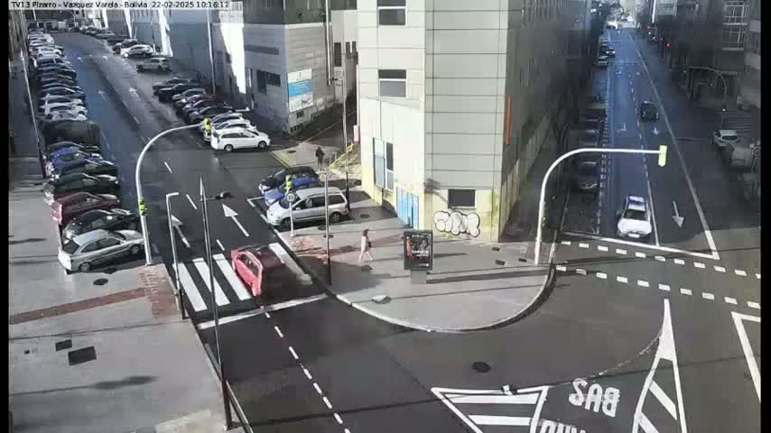 A sunny city street intersection shows parked cars lining both sides of the street and a few cars driving, with a pedestrian walking near a building.