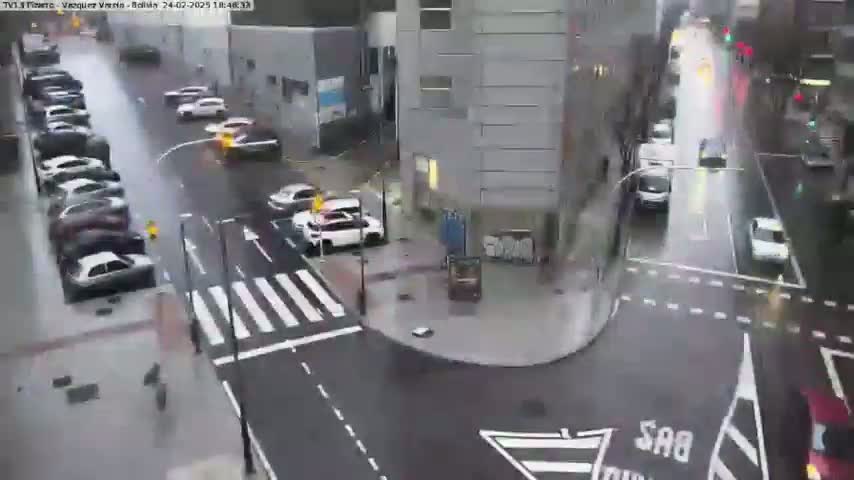 A rainy city street scene shows several cars parked along the side of a street and other cars driving through a wet intersection near a multi-story building.